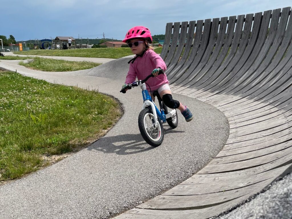kidsbikebalancebike A young girl with a pink helmet rides a balance bike at a pump track.