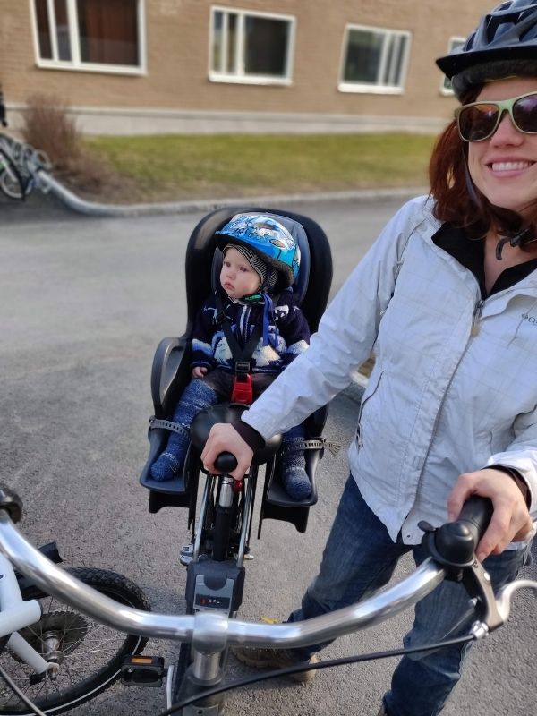 A woman in a white rain jacket and sunglasses holds up a bike with a baby in the bike seat, wearing a blue helmet and sweater. 
