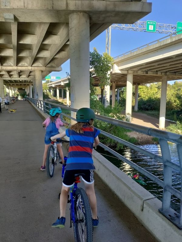 Two kids wearing helmets, one with overalls and one with a striped shirt and athletic shorts, are stopped on a bridge under a highway. They are looking down at the water, where you can barely see a kayaker. 