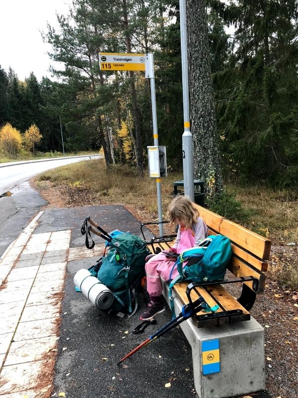 A girl in pink rain overalls sits on a bench reading at a bus stop. She is surrounded by backpacking bags, hiking poles, and sleeping mats. It's fall, the leaves are yellow, and it's wet outside. 