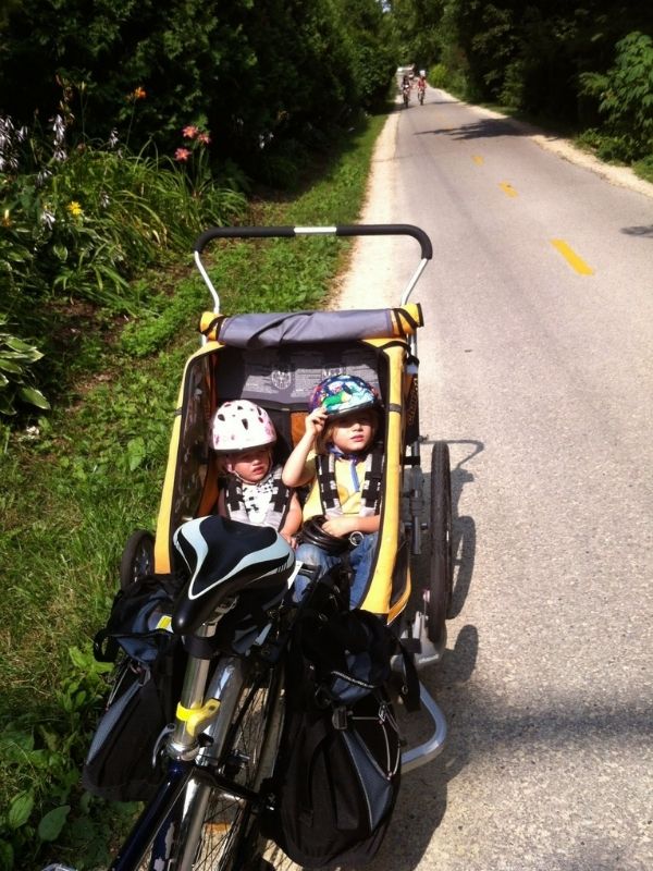 Two kids, each in helmets, sit inside a yellow bike trailer, they are stopped on a paved, divided bike path in the woods. 
