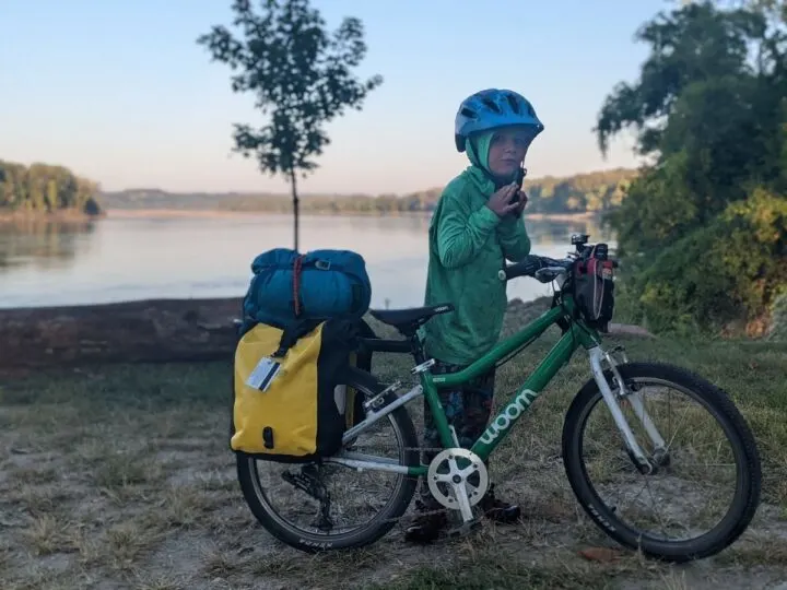 A young boy poses in front of the Missouri river with a bike loaded with camping gear.