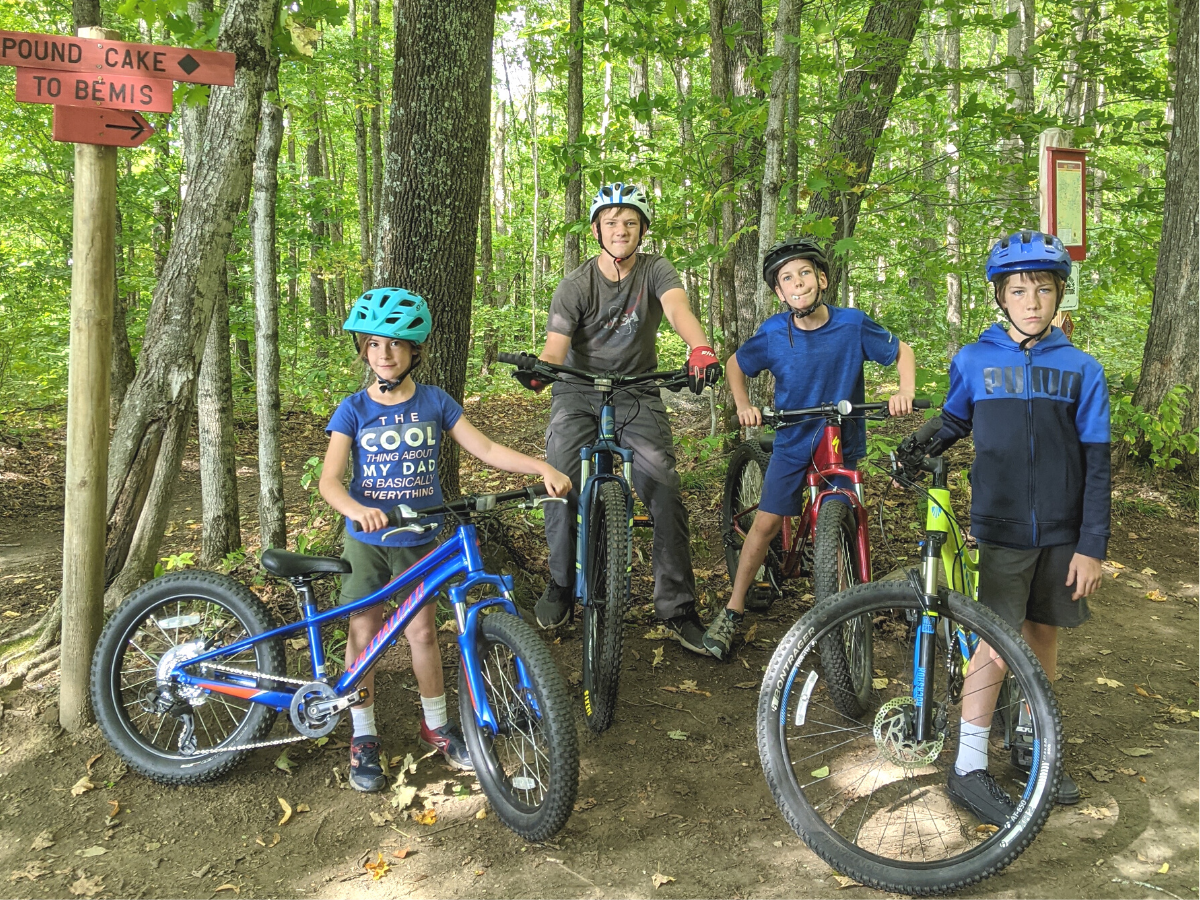 Four kids stand in the woods with mountain bikes wearing helmets. 