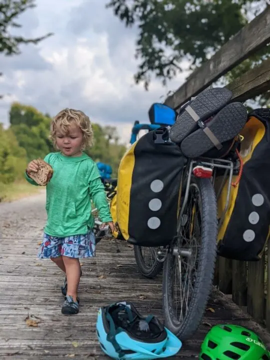 A sweaty toddler walks with a sandwich next to a loaded bike.