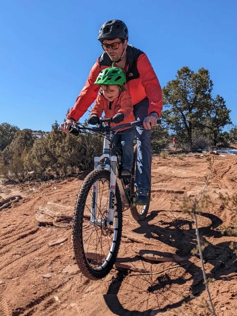 A small child rides on a bike with his dad over red dirt.
