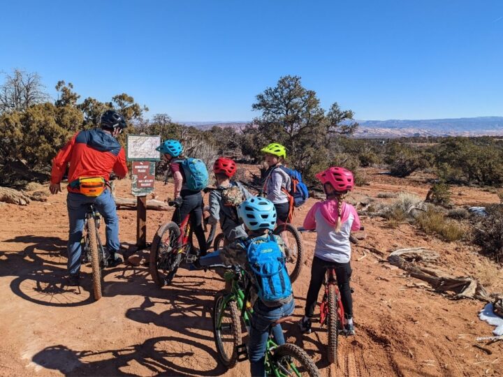 A dad studies a map on a trail surrounded by several kids on mountain bikes.