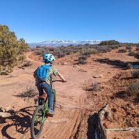A boy rides down a mountain bike trail with red rock and mountain views.