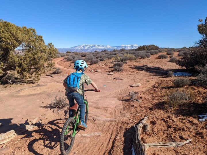 A boy rides down a mountain bike trail with red rock and mountain views.