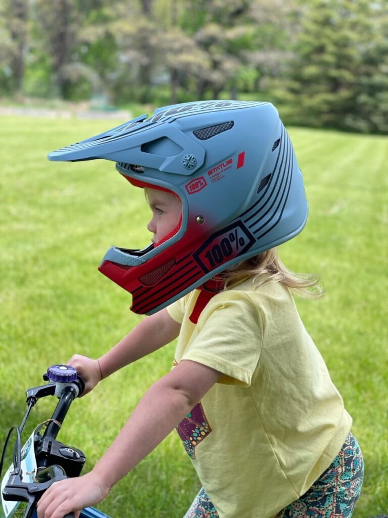 A young girl with a 100% Status Youth helmet on stands in a grassy yard.