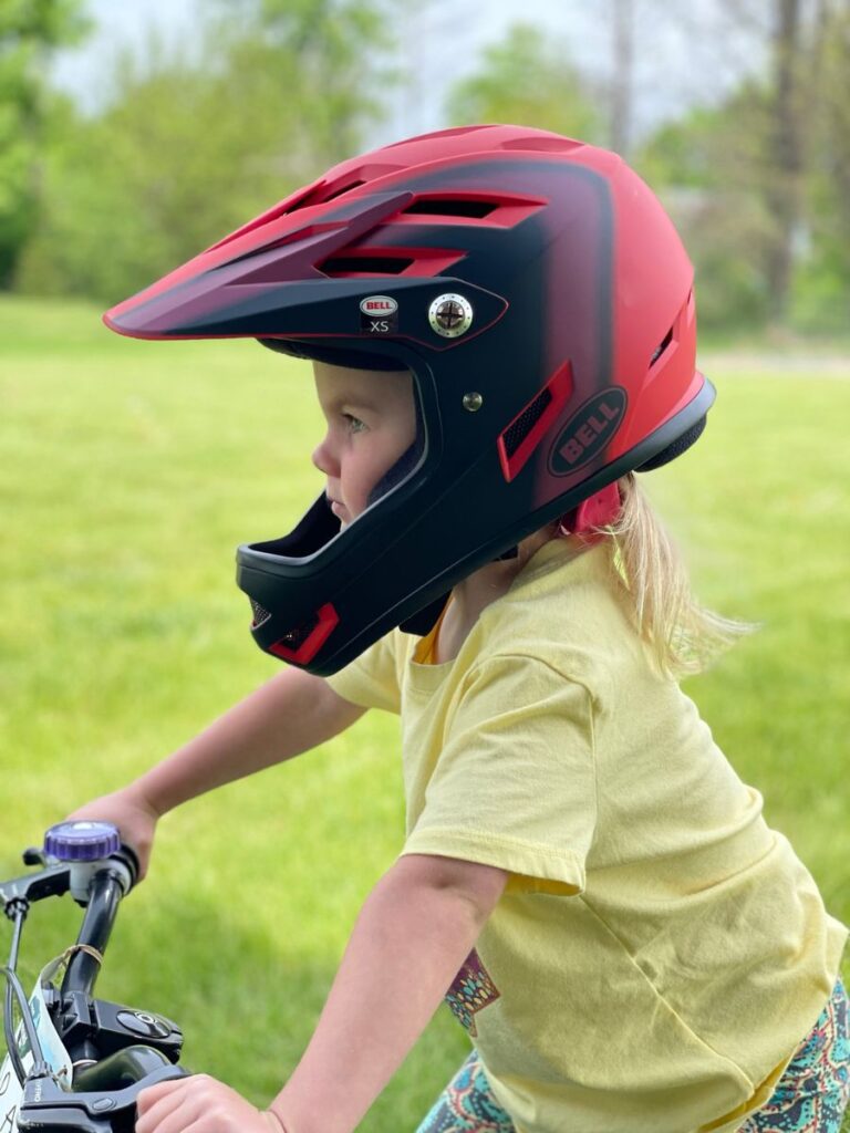 A young girl with a Bell Sanction helmet on stands in a grassy yard.