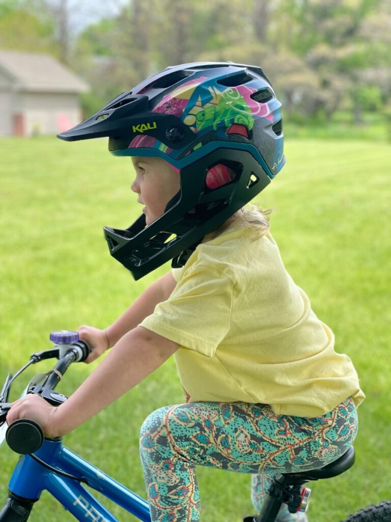 A young girl with a Kali Maya Full Face helmet on stands in a grassy yard.