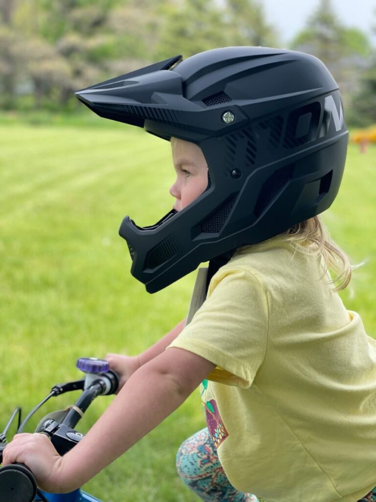 A young girl with a Mongoose Title helmet on stands in a grassy yard.