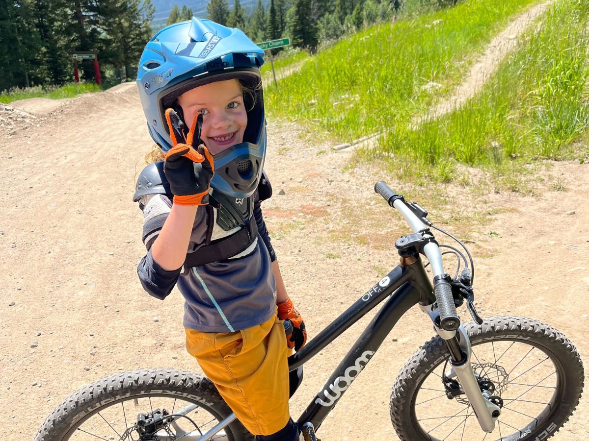A young girl wearing a 7iDP M1 helmet stands over her bike on a gravel trail giving a peace sign.