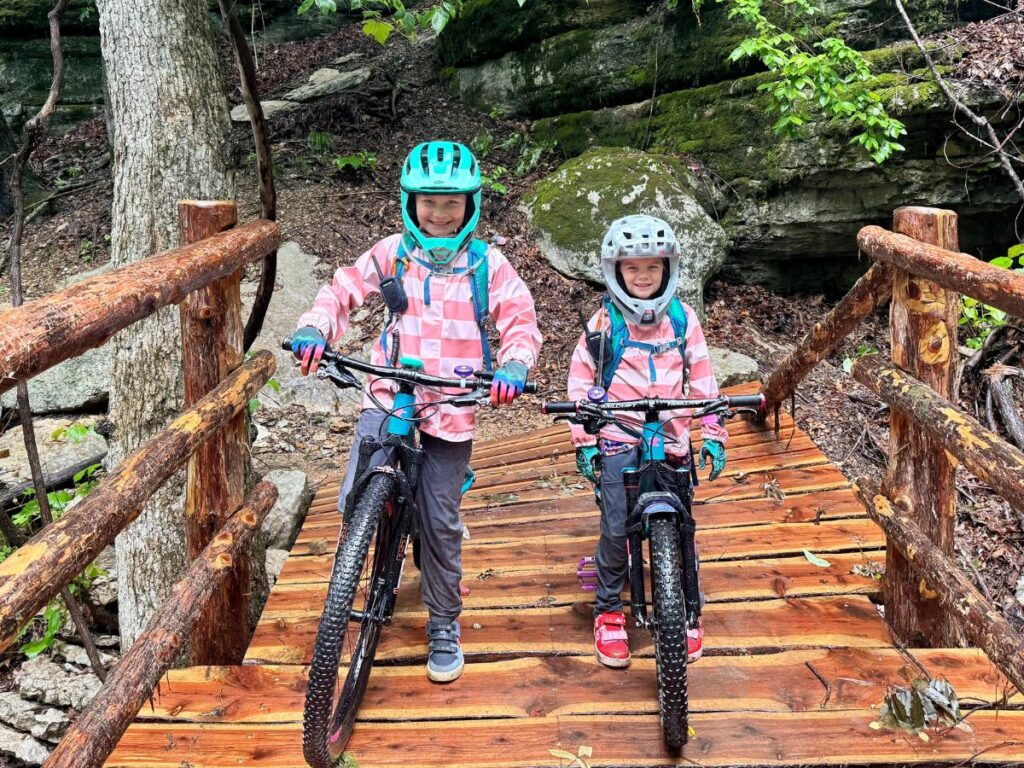 Two girls stand over their bikes on on a wet bridge. They are wearing full face helmets and rain jackets.