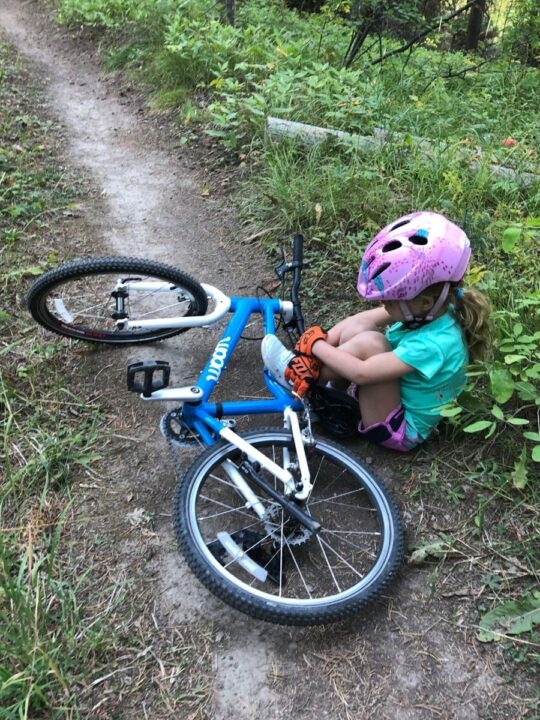 photo of a child wearing a bike helmet and bike gloves sitting on the ground next to her bike. She needs bike gloves for kids.
