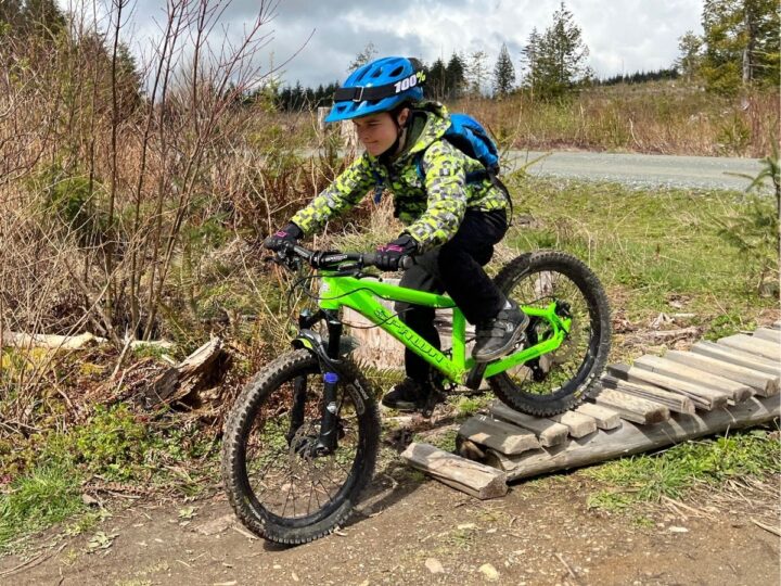 photo of a child wearing a bike helmet and bike gloves, riding a mountain bike across a wooden feature
