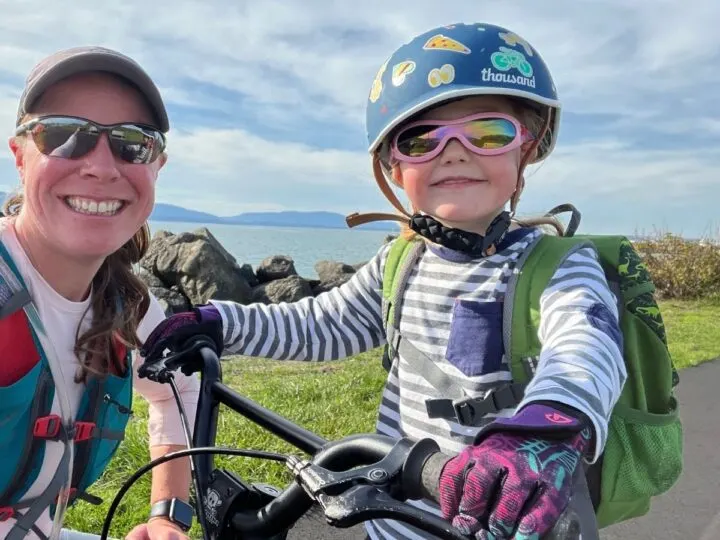 photo of a woman crouching down next to her child, who is riding her bike and wearing a bike helmet, bike gloves, and sunglasses