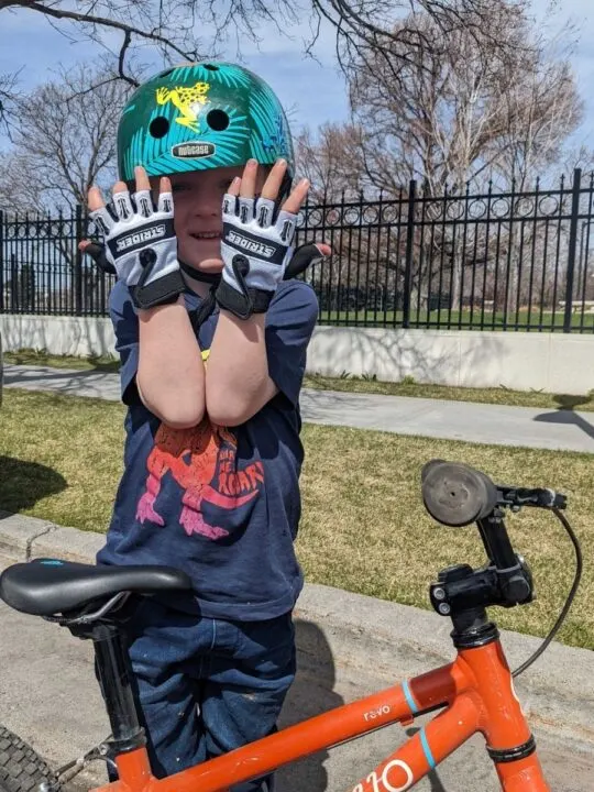 photo of a child standing next to his bike wearing Strider half-finger bike gloves and a bike helmet