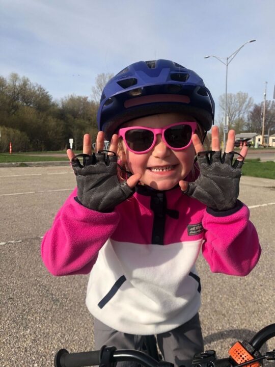 photo of a child holding up her hands wearing a bike helmet, bike gloves, and sunglasses