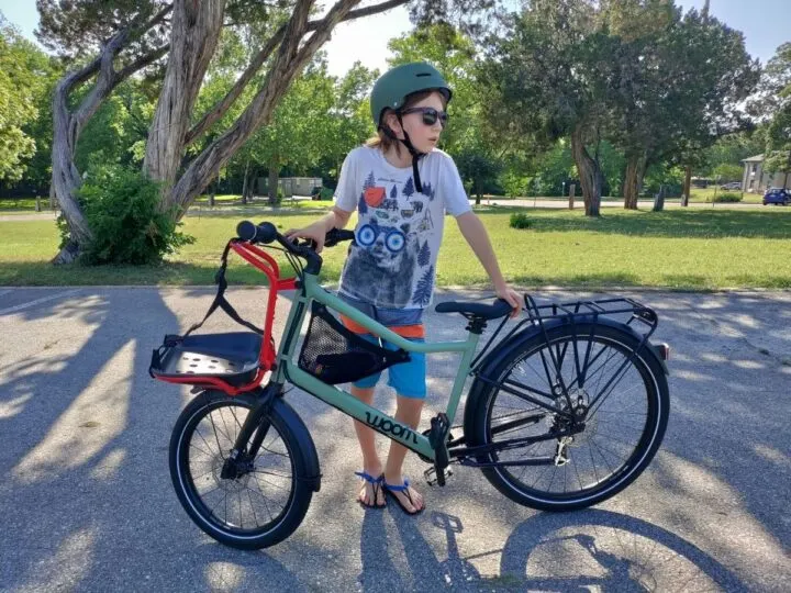 A pre-teen kid stands next to his bike, looking very cool in sunglasses and sandals. Woom now review. 