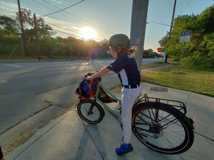A kid in a green helmet is stopped at an intersection on a bike. The sun is setting. 