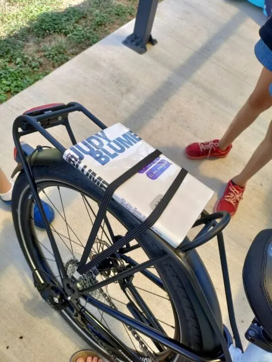 A Judy Blume novel is secured onto a kid's bike rack. 