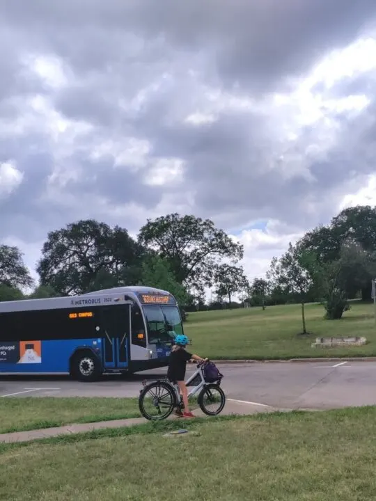A kid in a blue helmet and black shirt is stopped on a sidewalk, by a road. A blue city bus is driving past. 