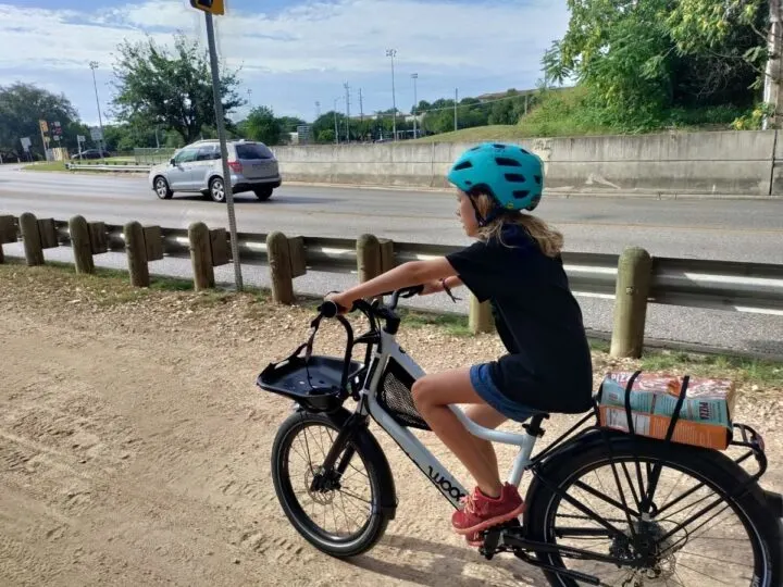 A long-haired kid in a helmet rides a bike on a dirt trail next to a main road. There's a barrier in between the trail and road. 
