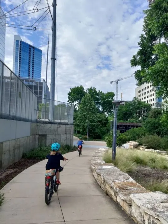 Two kids on bikes ride through downtown Austin, TX on a path. 
