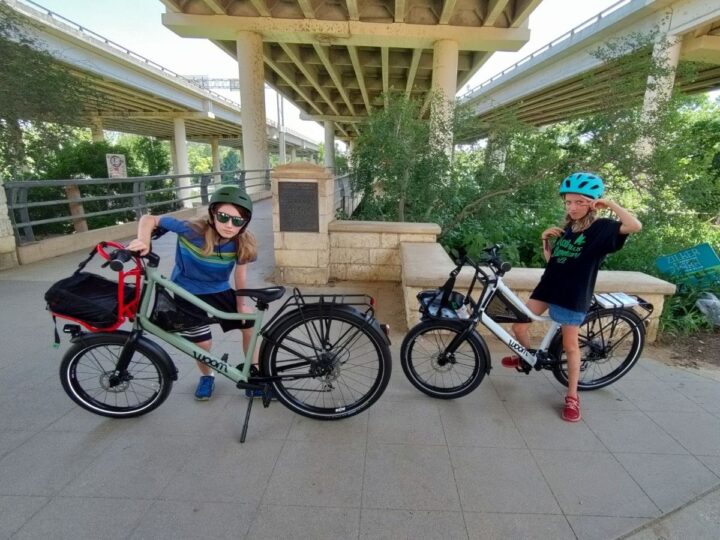 Two long-haired blonde kids in helmets and t-shirts pose with their bikes on a pedestrian passage under an overpass. 