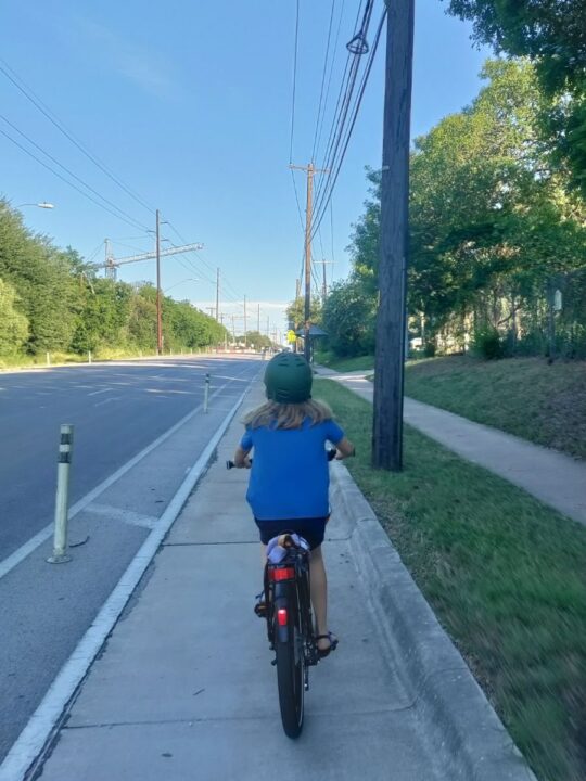 A long-haired kid in green helmet and blue shirt rides a bike down a bike lane next to a sidewalk, electric poles, and four-lane road. 