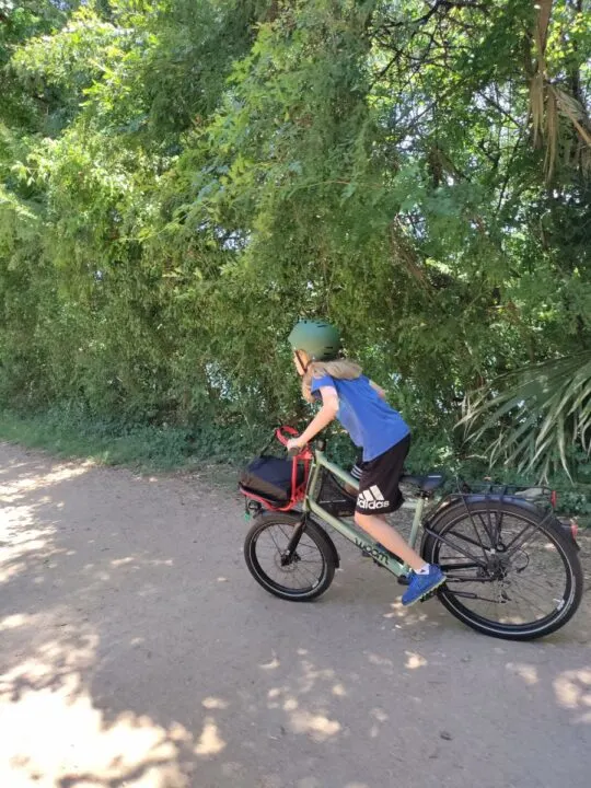 A long-haired kid in a blue shirt rides a bike on a dirt trail with trees in the background. He's standing up and pedaling aggressively. 