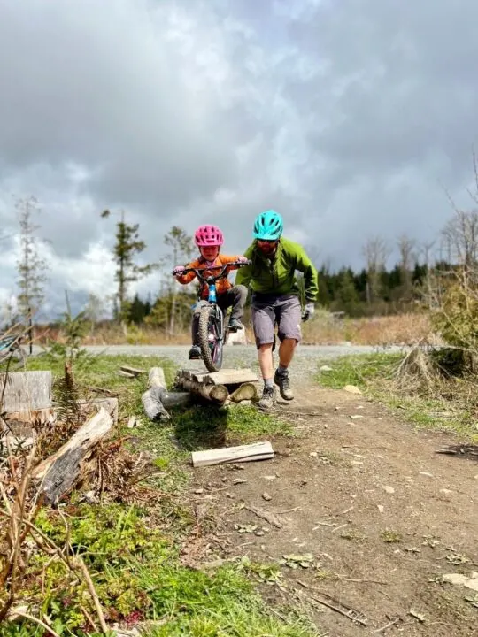 Photo of a child riding a mountain bike across a wooden see-saw feature while being helped by an adult