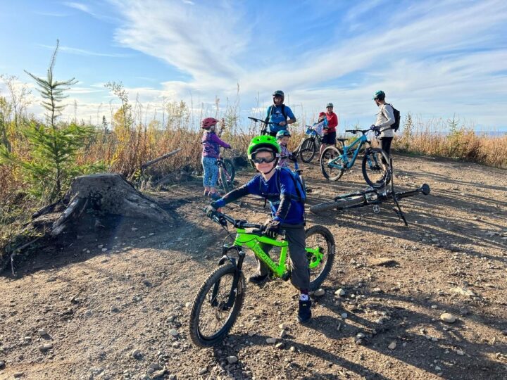 Photo of a child standing over his bike in a dirt area, with several other adults and children with bikes in the background