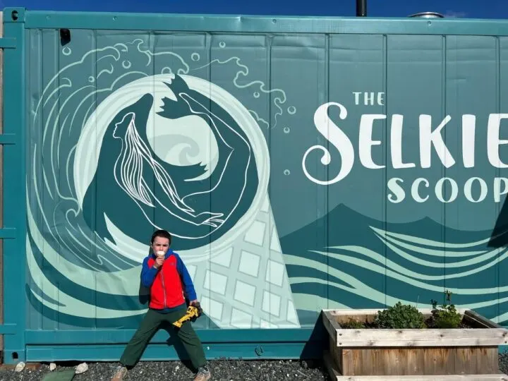 Photo of a child holding an ice cream cone standing in front of a shipping container with a mural and the words "The Selkie Scoop"