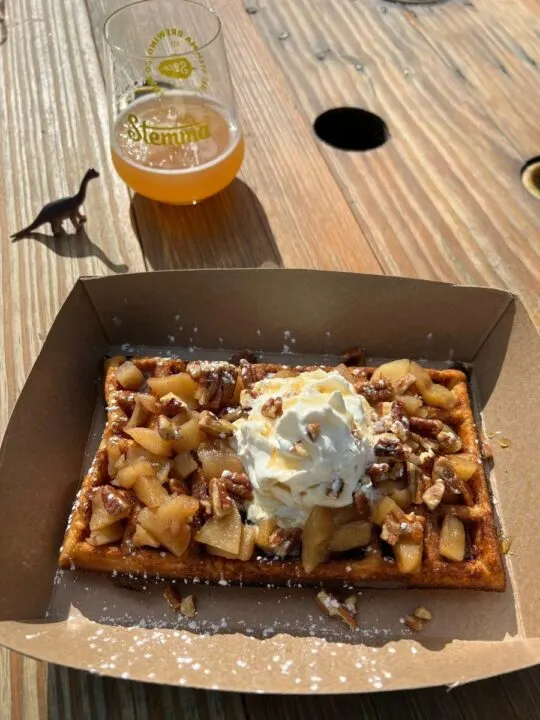 Photo of a waffle topped with apples, nuts, and whipped cream sitting on a table alongside a toy dinosaur and an orange-colored beverage