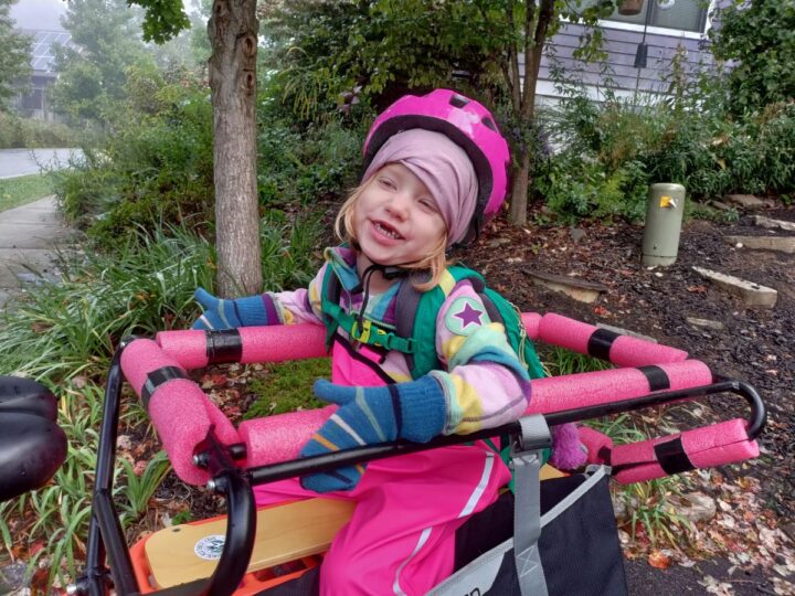 A little girl in a rainbow fleece and pink rainpants sits on the back of a longtail cargo bike