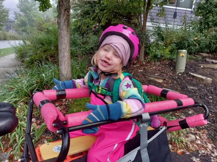 A little girl in a rainbow fleece and pink rainpants sits on the back of a longtail cargo bike