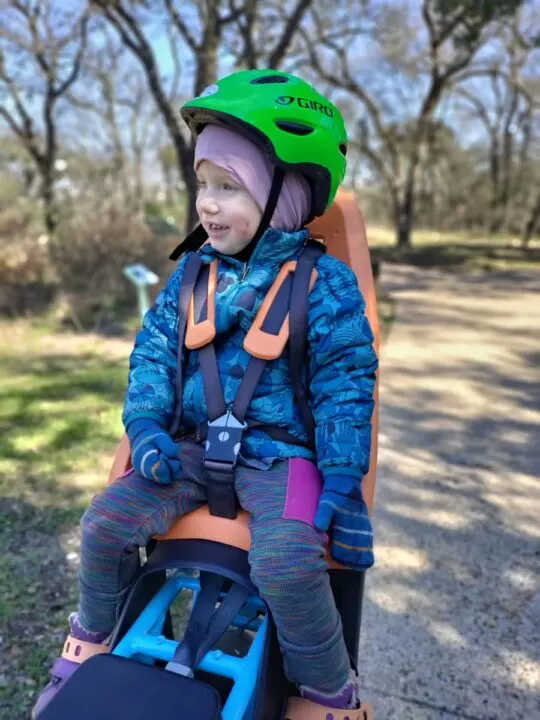 A child sits in an orange bike seat wearing a thick down coat, mittens, and rainbow pants