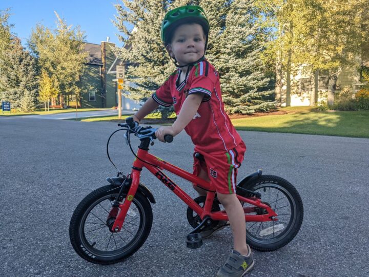 A boy dressed in red poses atop a tiny red pedal bike. 
