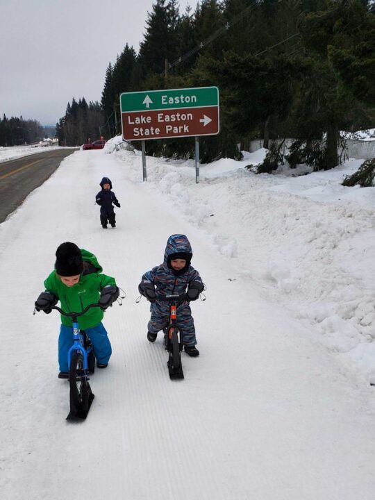 A kid in a green jacket and one in a multicolored snow suit bike on kickbikes through a snowy path