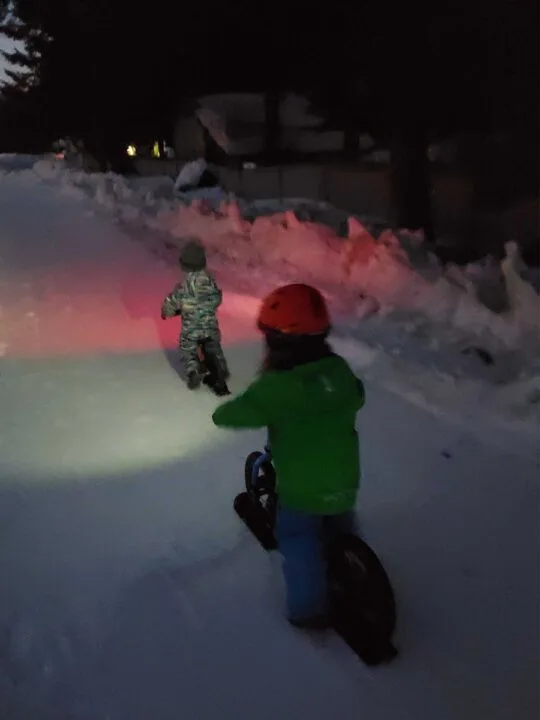 Two kids on bikes have lights on them and are biking in the dark.