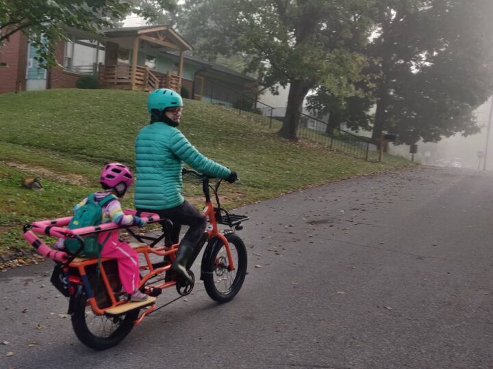 A woman in a teal jacket rides a longtail bike with a kid on the back, she has a teal backpack and pink rainpants. 