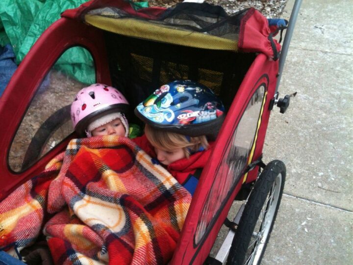A kid and a baby sit in a red bike trailer, snuggled up in a plaid wool blanket