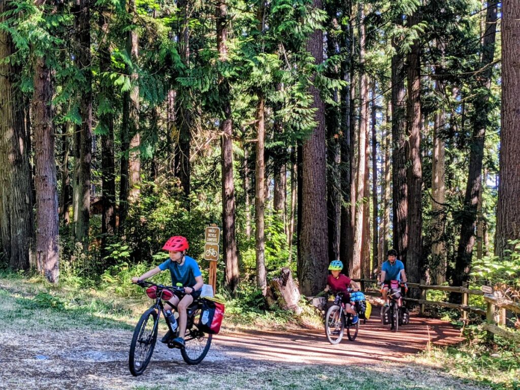 A family rides loaded touring bikes out of a wooded trail.