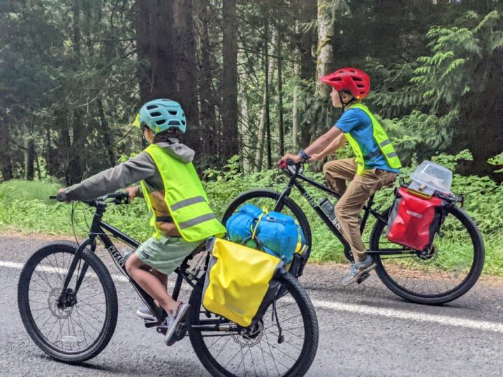 Two boys bike along a road wearing bright yellow vests.