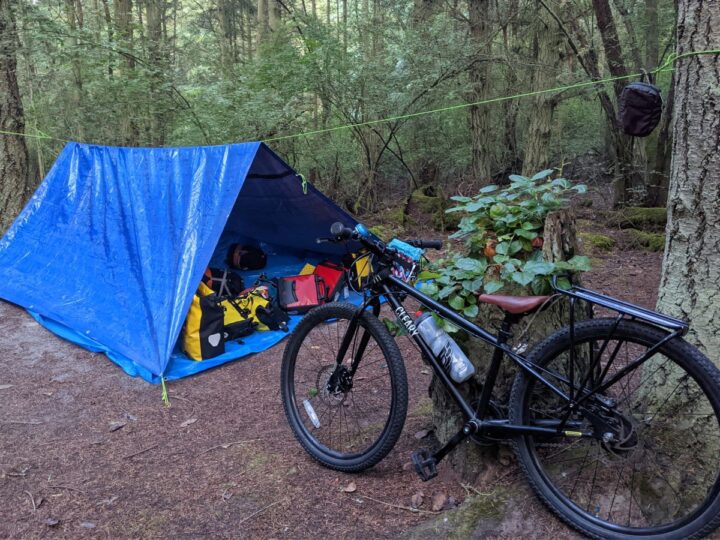 An A-frame tarp is set up in the trees with a bike in the foreground.