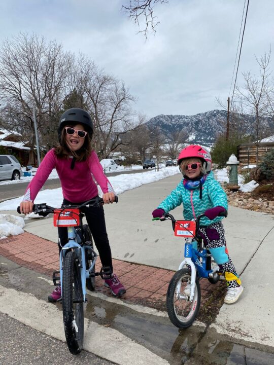 Two girls stand atop their bikes, next to a curb. It appears to be cold, with one girl wearing a coat and gloves. They are also wearing sunglasses. There are mountains in the background. 