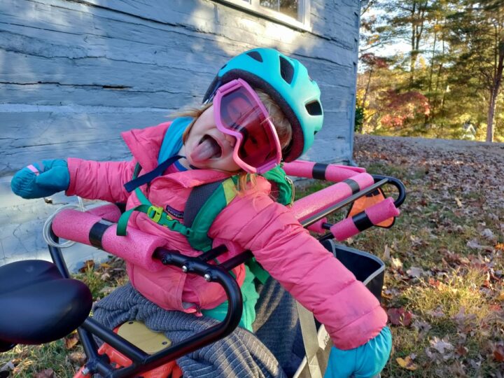 A little girl on a longtail bike wears a pink jacket, pink ski goggles, and a blue helmet. 