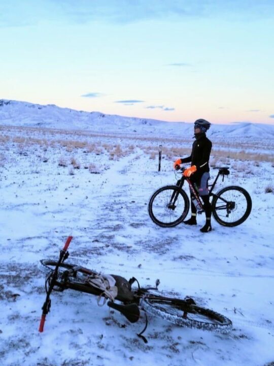 An adult stands with a bike in the snow, there are mountains in the distance and a second bike in the foreground.